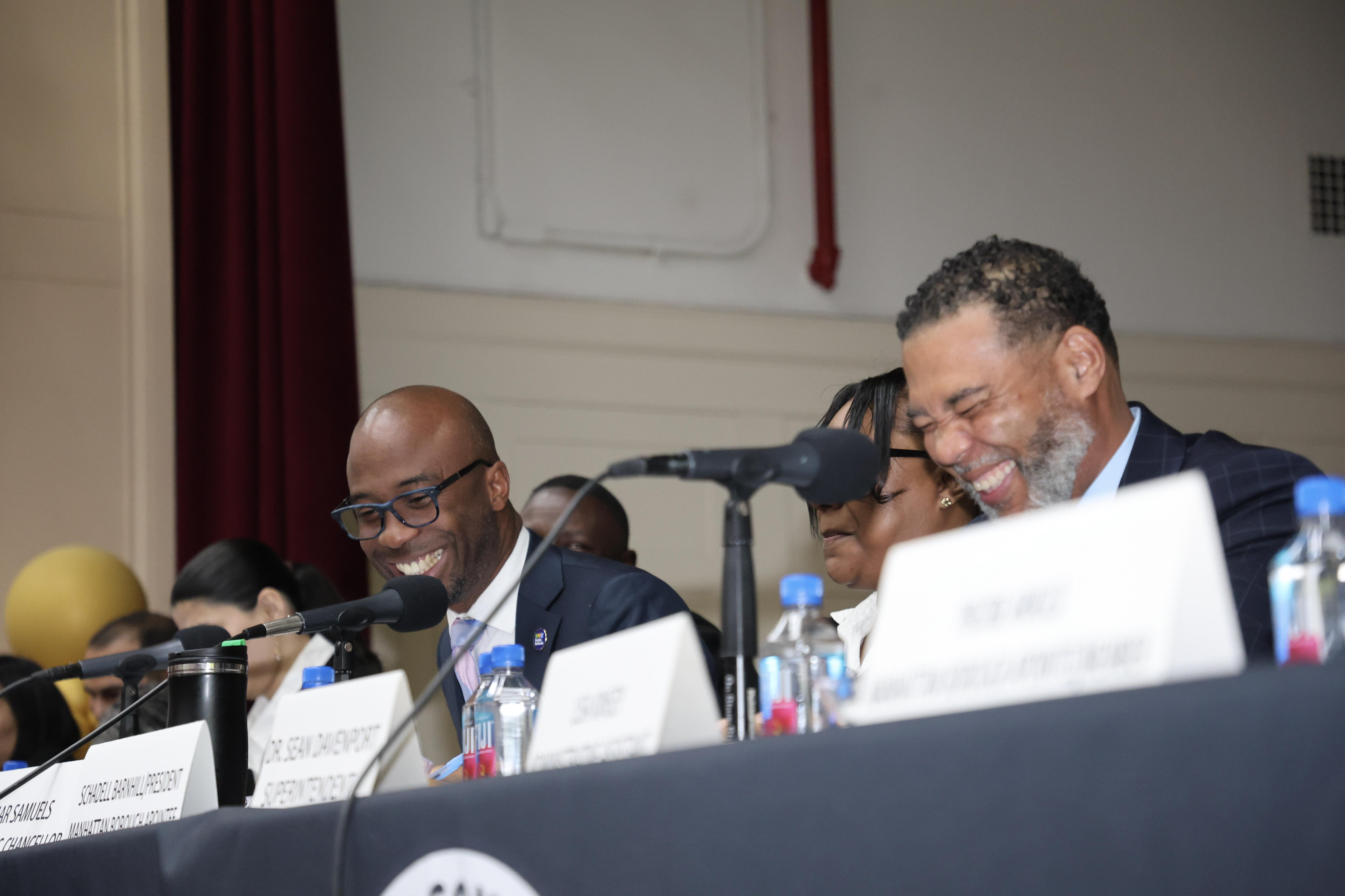 Two men laughing and speaking at a meeting table with water bottles.