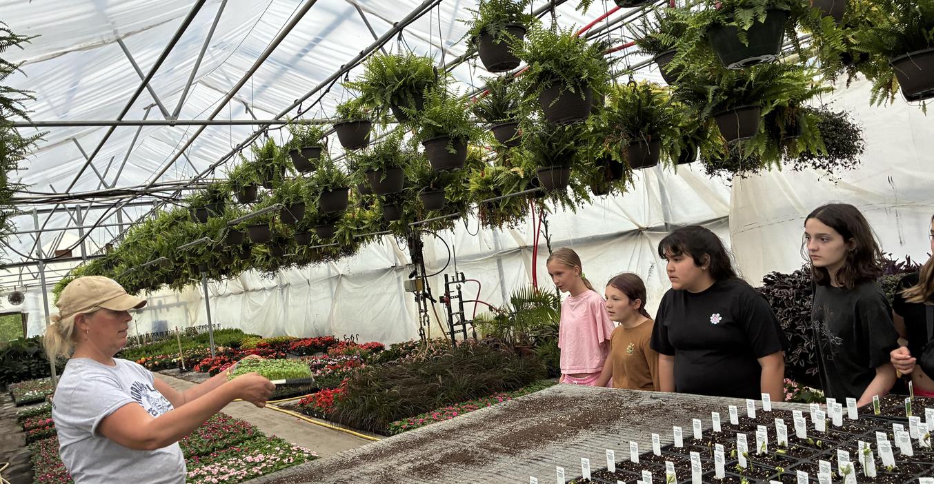 A group of children observes a woman in a greenhouse filled with hanging plants.