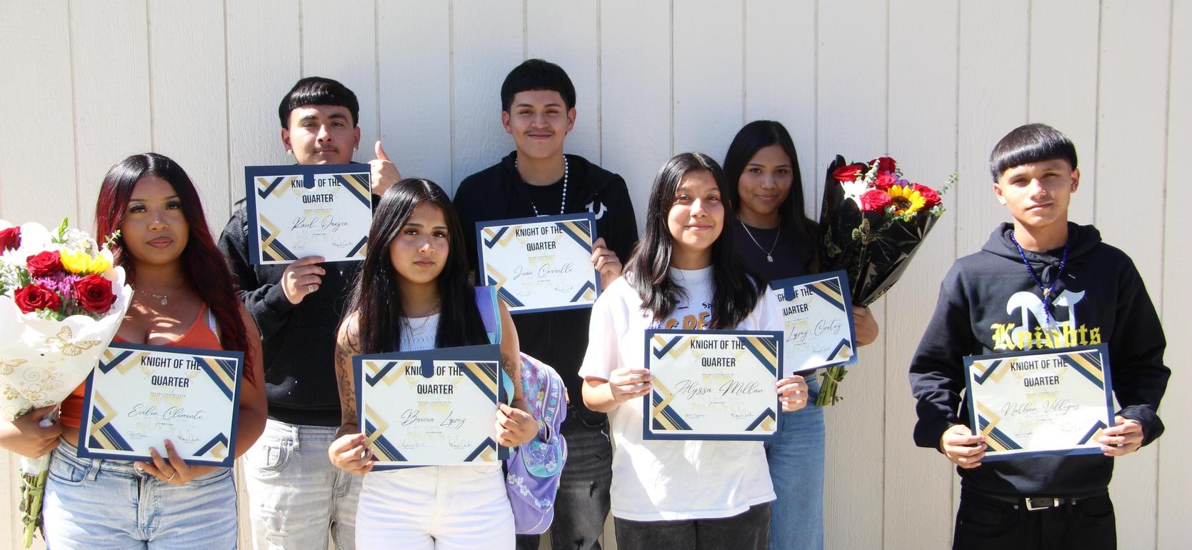 Group of students holding certificates and flowers, smiling in front of a wall.