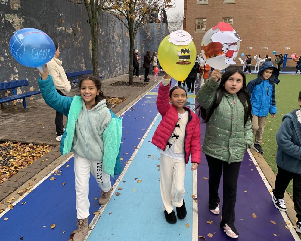 Three children in a school parade walking on a colorful path, each holding a balloon.