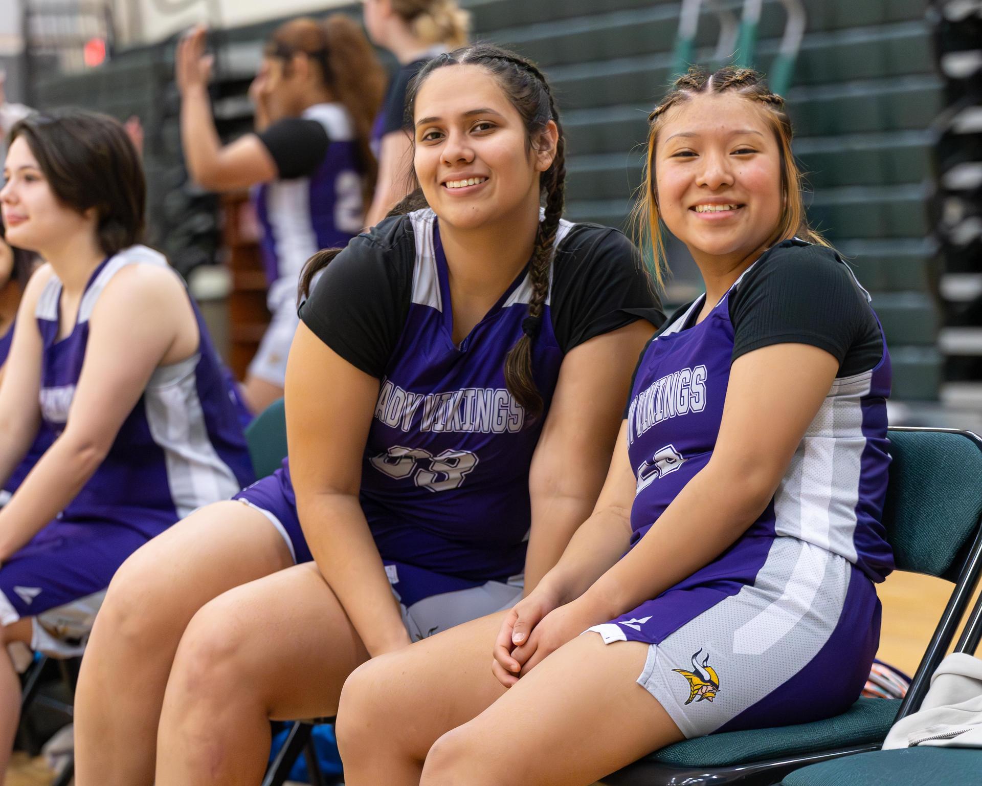 Two varsity girls basketball players posing for a picture on the sidelines during a game.