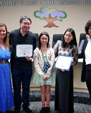 Five individuals, including a child, hold certificates at a school board meeting.