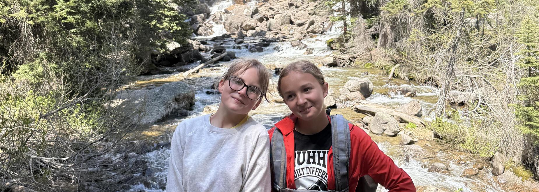Two girls stand together with a waterfall in the background.