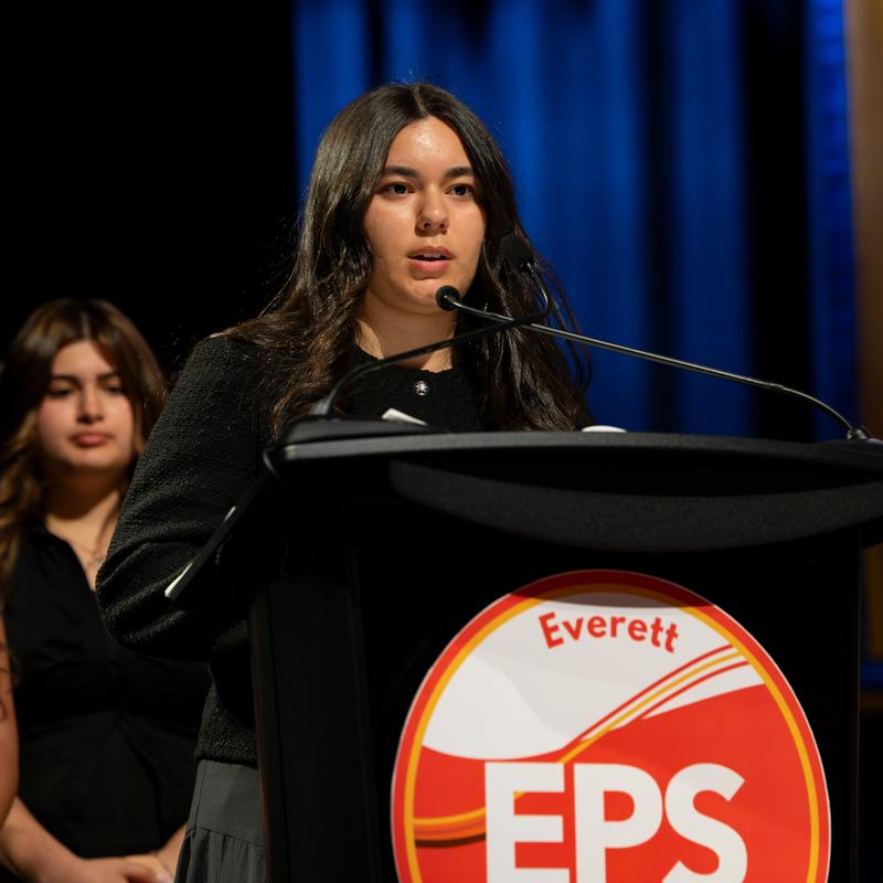 A young woman speaks at a podium, with another person in the background.