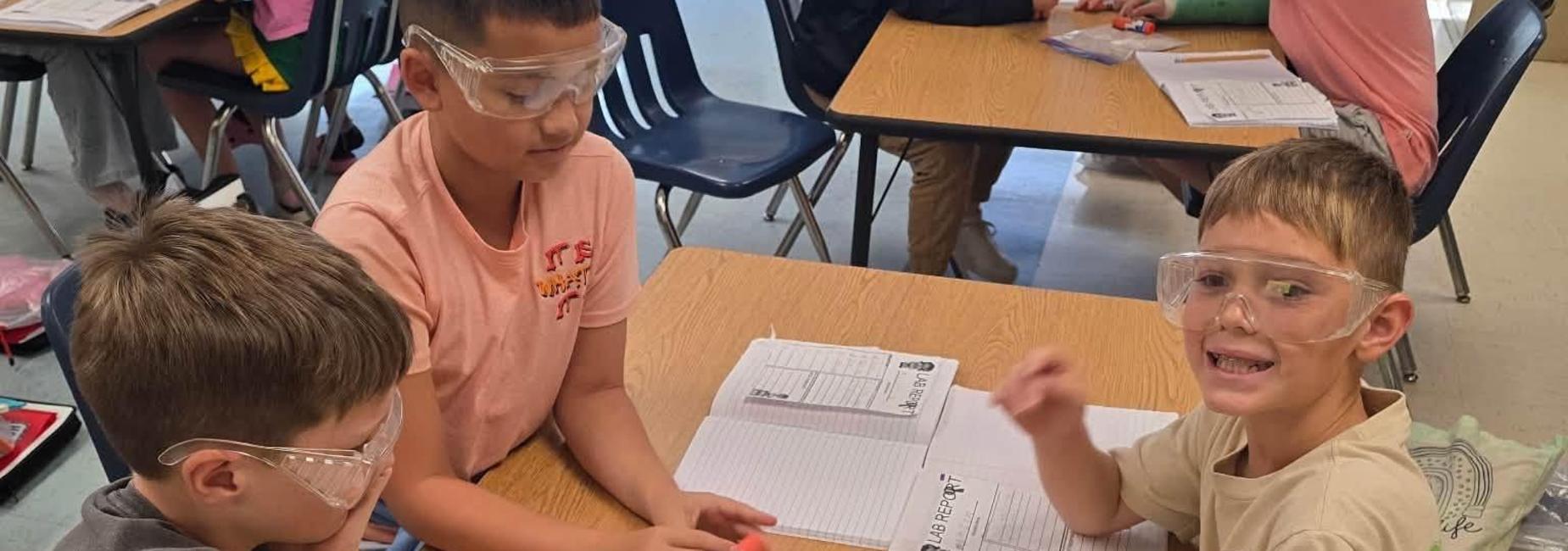 Two children working together at a table with safety goggles and a bag of materials.