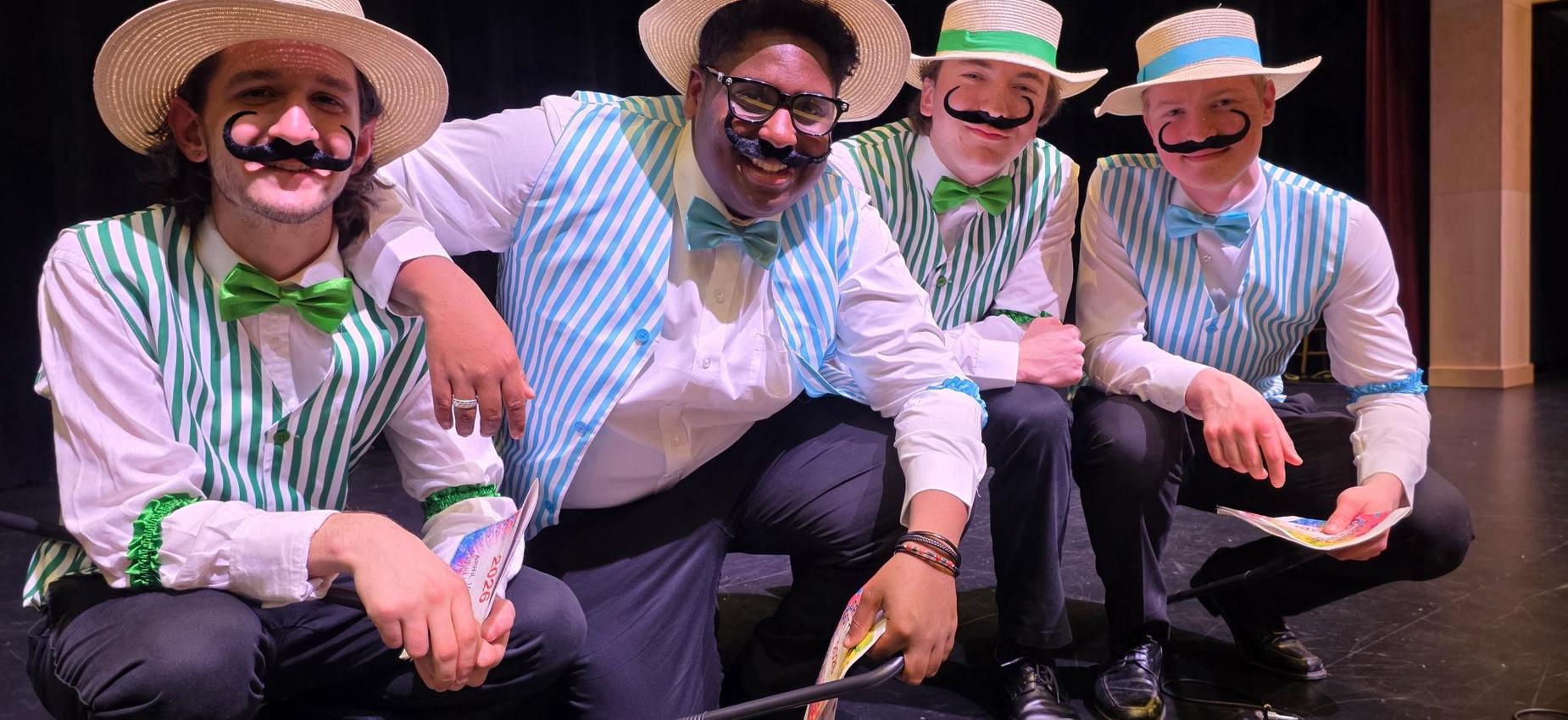 Four performers in striped vests and hats pose playfully with mustaches.
