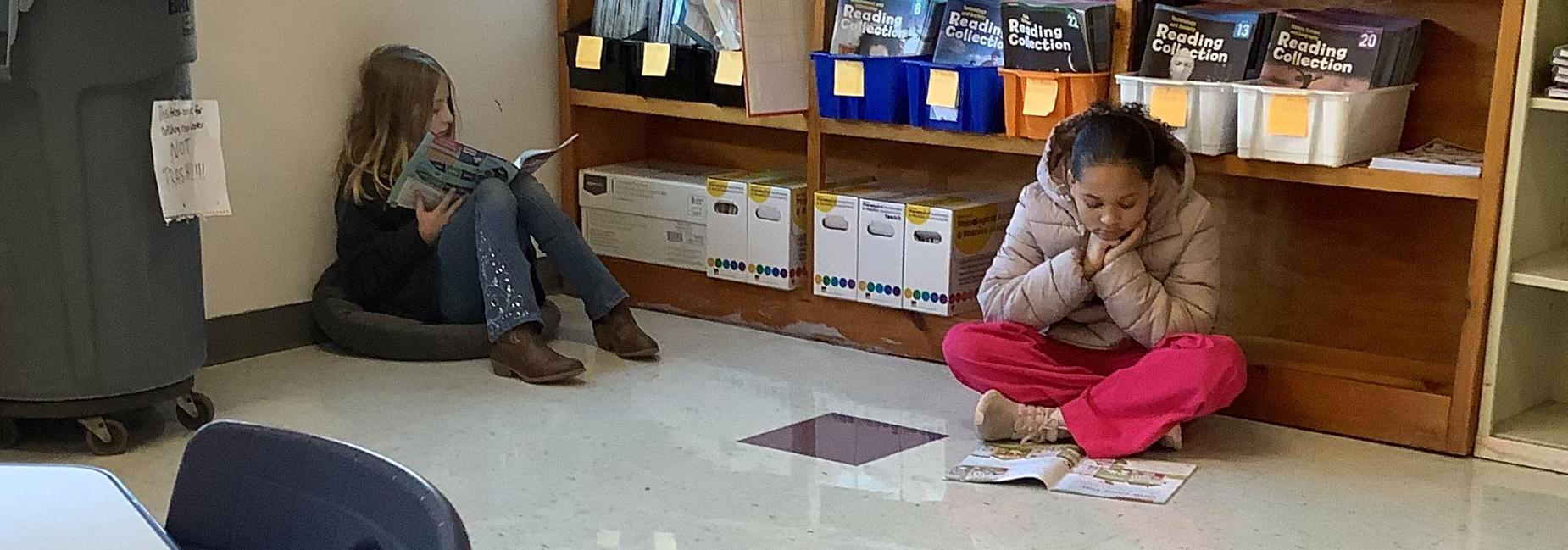 Two children reading on the floor in a classroom with boxes and shelves in the background.