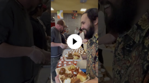 Adult smiling as he receives a full Thanksgiving-style meal from staff serving trays of food in a busy cafeteria.