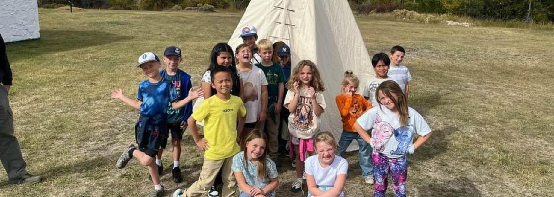 Group of children posing cheerfully in front of a teepee on a grassy field.