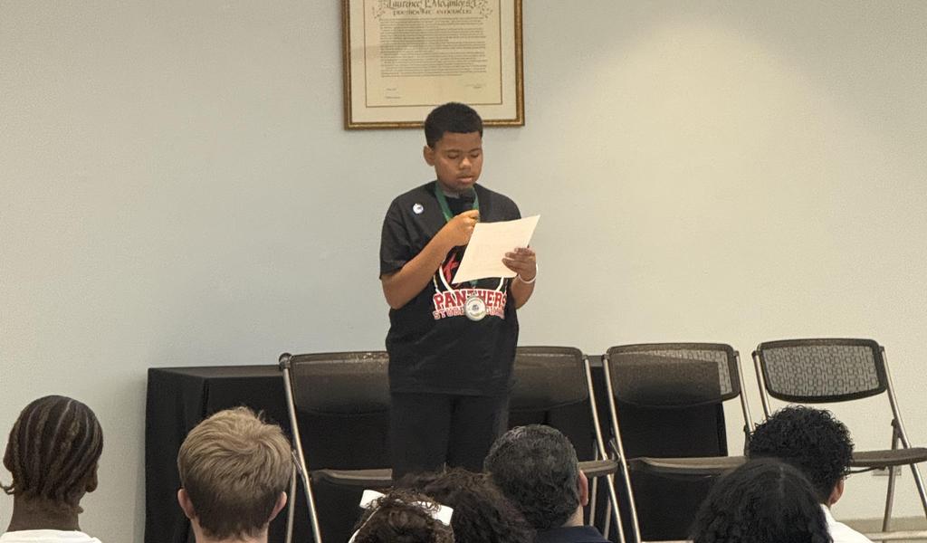 A young boy reads a speech at a podium, with an audience seated behind him.