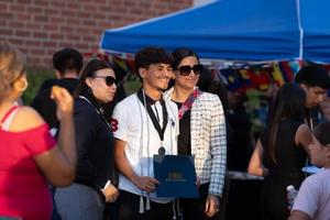 A student proudly poses with family members outside, holding his PUSD Seal of Biliteracy Award folder while wearing a medal and cord, with flags and celebration decorations in the background.