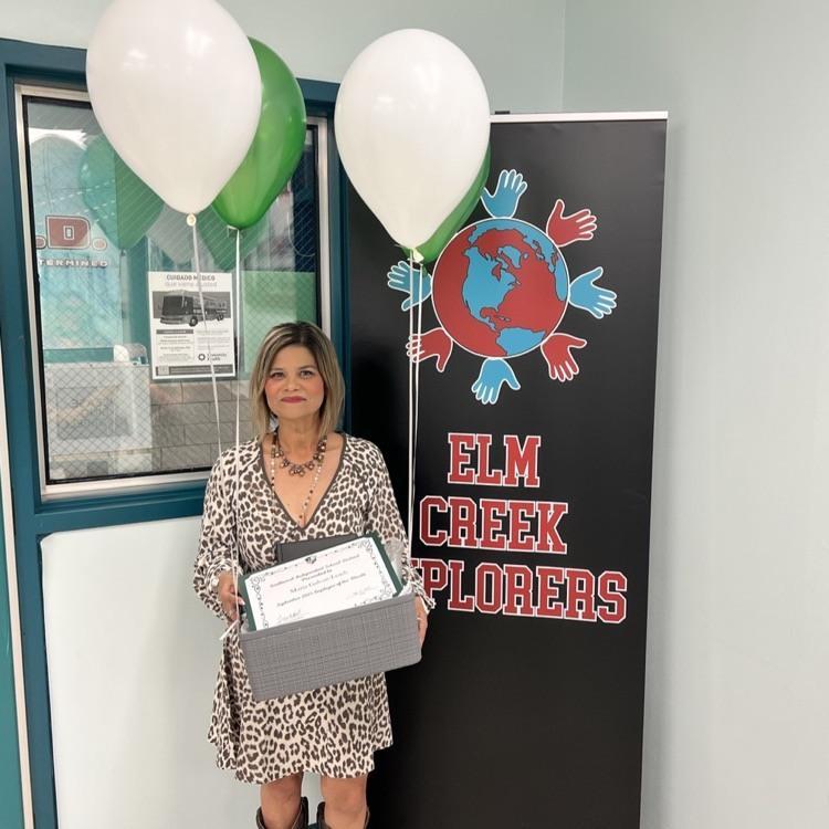 Woman wearing dress and boots, standing in front of black, maroon, and teal Elm Creek sign, holding basket of goodies and green and white balloons