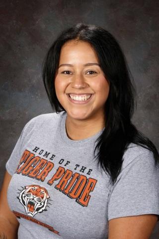 Smiling teacher with dark hair wearing an orange shirt against a gray backdrop.