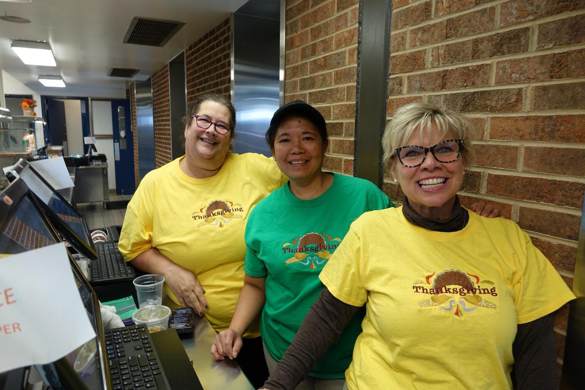 Three staff members in colorful shirts pose in a cafeteria setting.