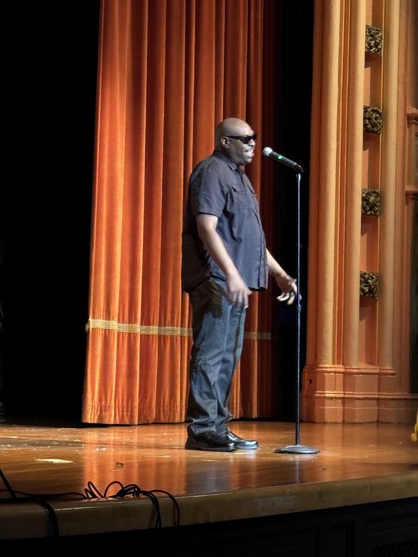 Man standing at a microphone on stage, wearing sunglasses and black clothing.