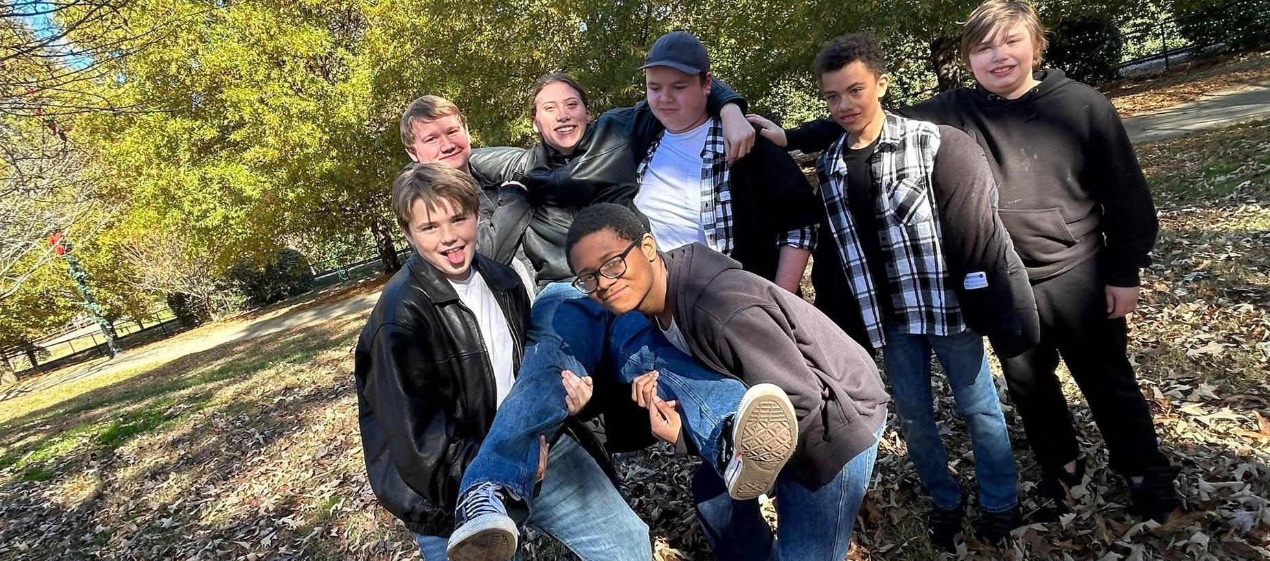 A group of seven smiling teenagers poses together in a park, surrounded by autumn leaves.