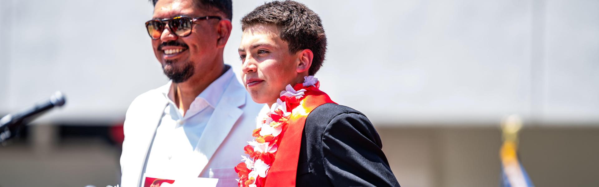 A young graduate wearing a lei smiles next to a principal Nunez