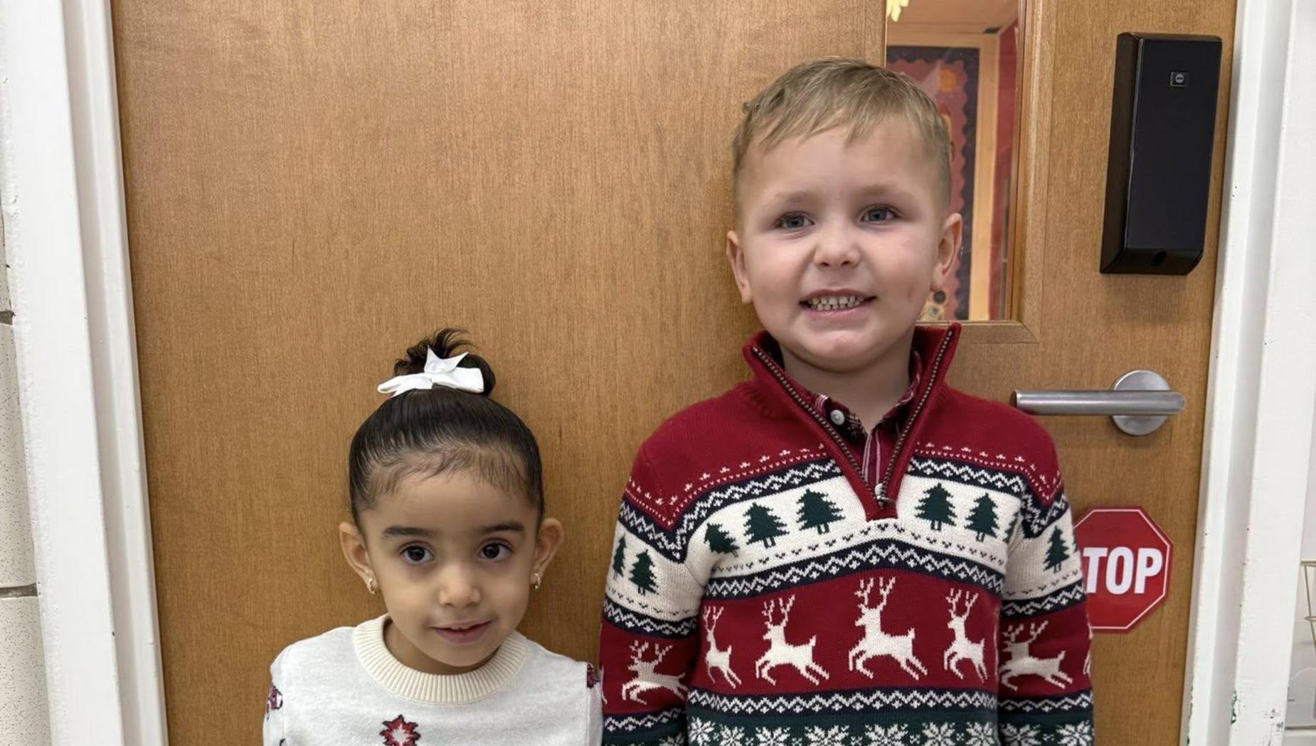 Two children in festive sweaters posing in front of a wooden door.