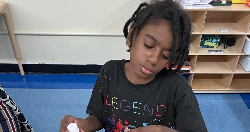 A child applying glue to colorful paper strips on a yellow background during a craft activity.
