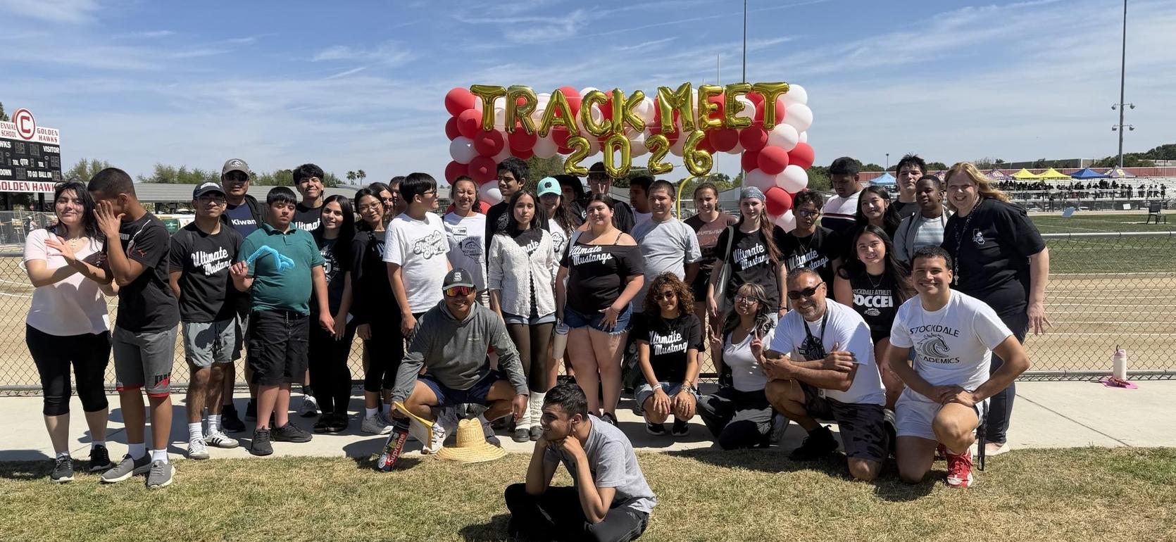 Large group of attendees at a track meet under a balloon arch.