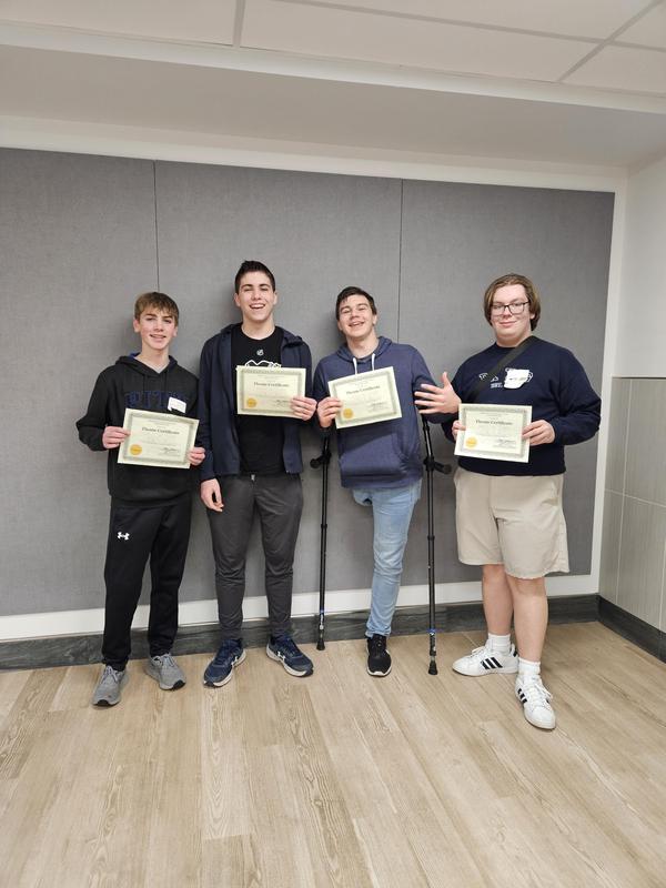 four boys smiling holding certificates leaning on gray wall