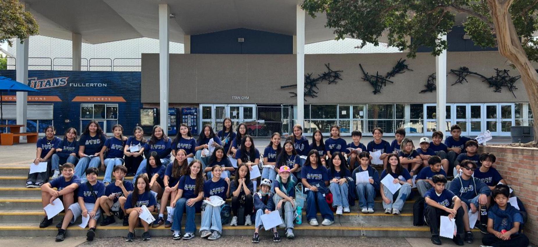 Group of students in matching blue shirts sitting on steps outside a building.