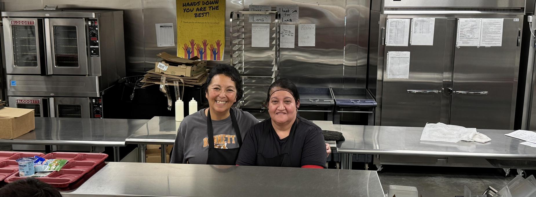 Two women smile at the camera in a kitchen setting with stainless steel equipment.