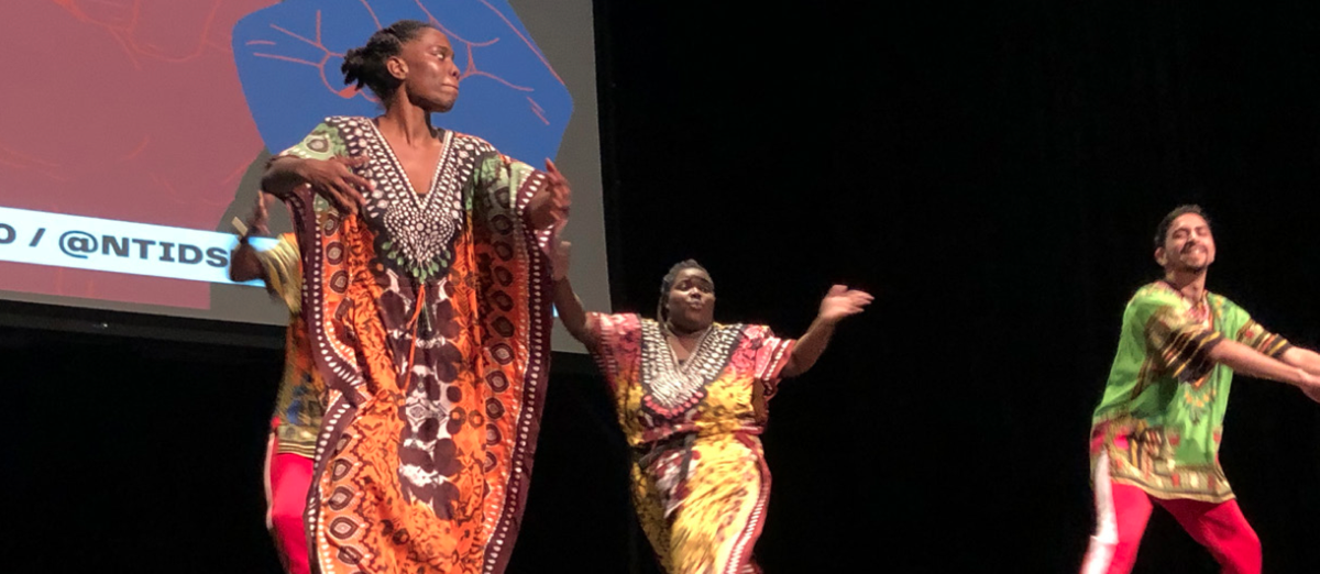 Performers from the Sunshine 2.0 group during a vibrant Black History Month celebration. Three dancers wearing colorful traditional African-inspired attire passionately perform on stage, expressing joy and energy through their movements. A projected background includes the NTID logo, adding context to the dynamic cultural performance.