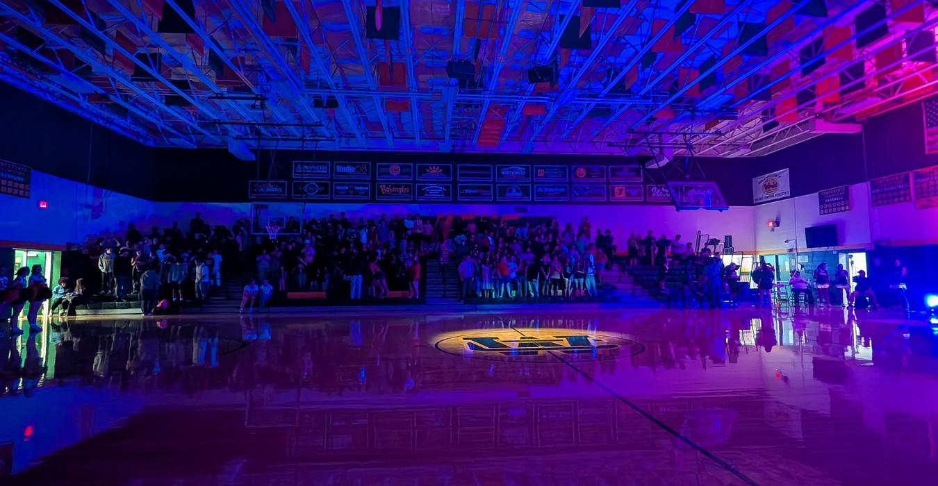 A dimly lit gymnasium with a large crowd, showcasing colorful banners and a gleaming floor.