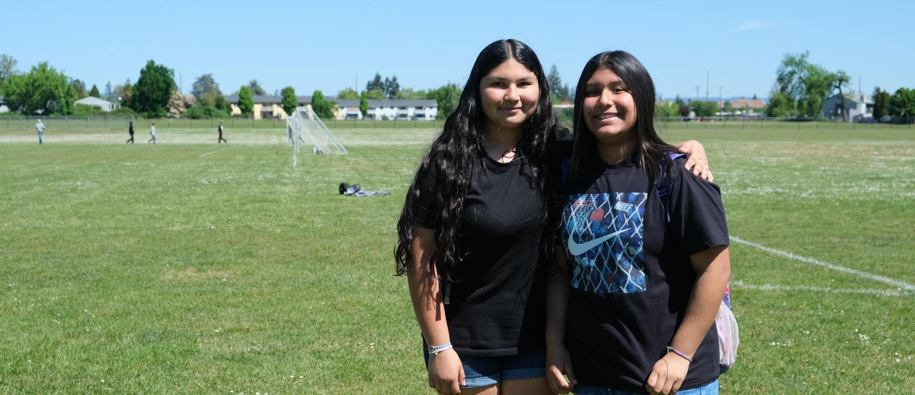 Two girls smiling together on a sunny field, wearing matching black shirts.