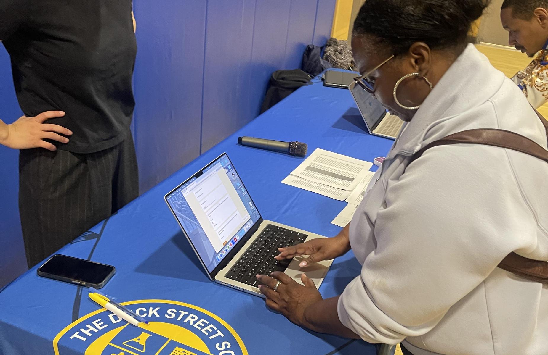 A woman types on a laptop at a table with various items around.