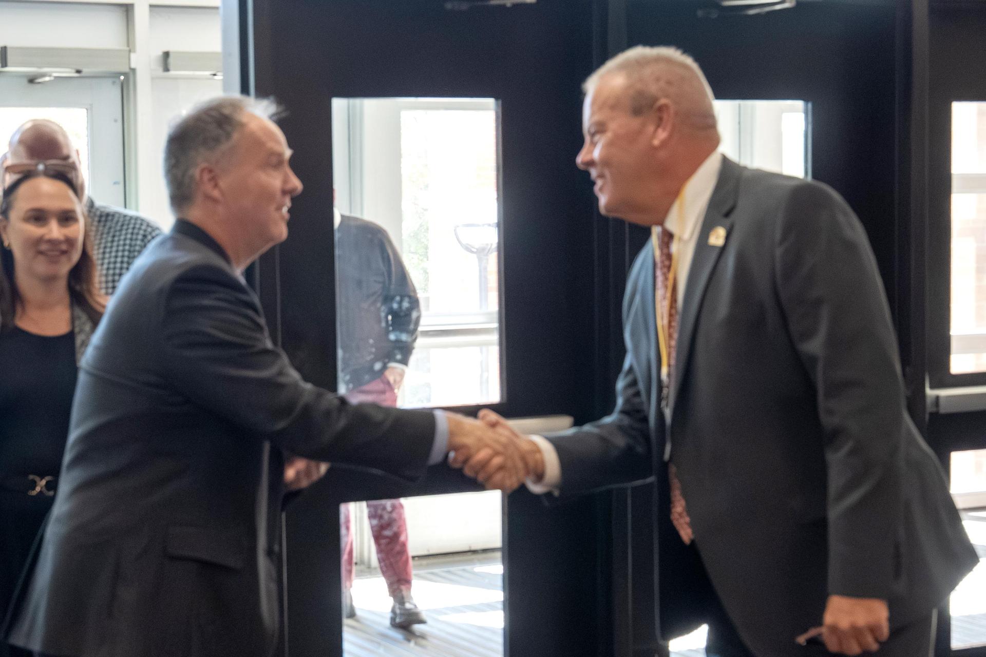 Two men shaking hands warmly in an indoor setting, with people in the background.