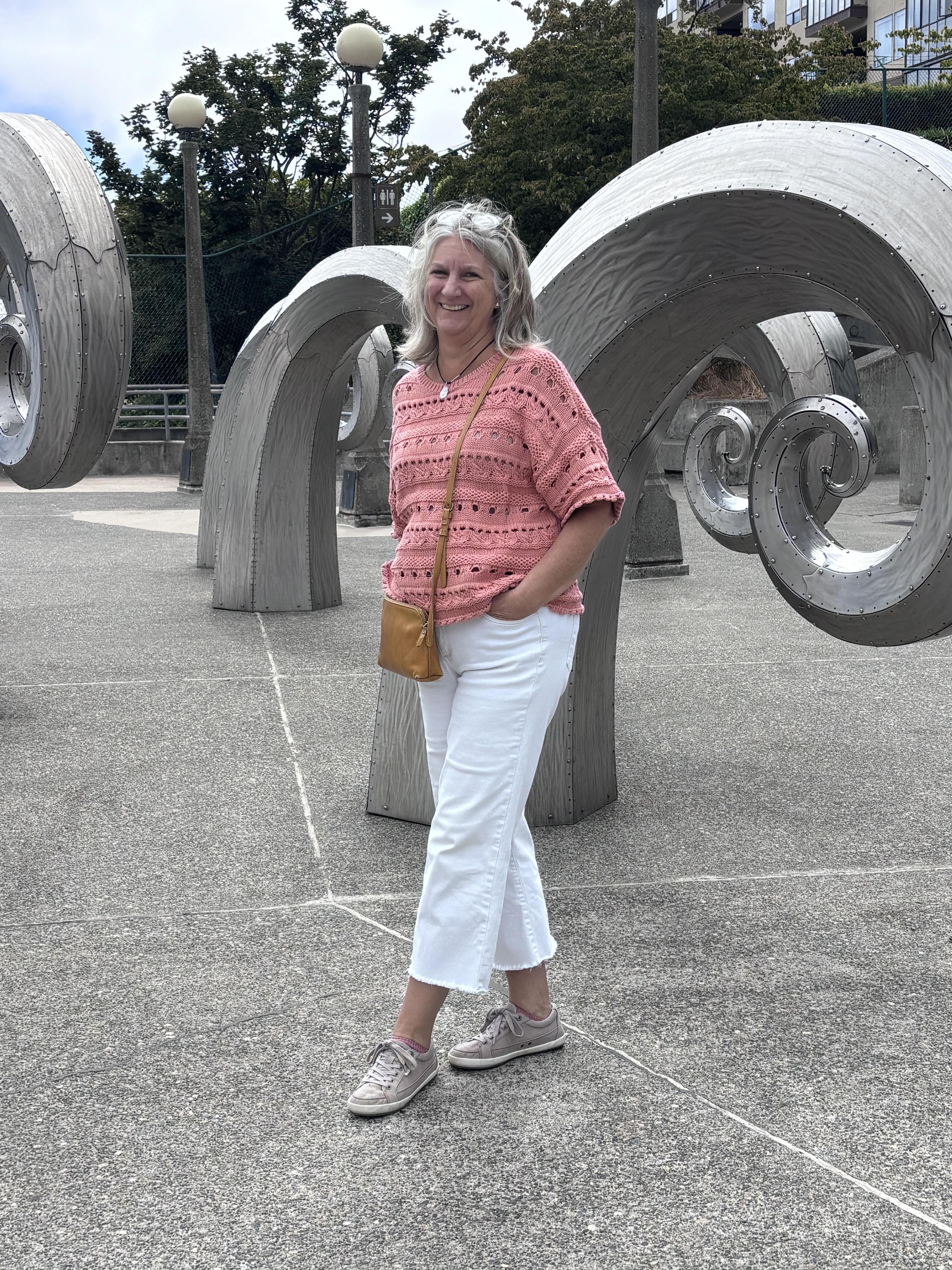 Woman in a pink sweater stands among curved wooden sculptures in an outdoor setting.