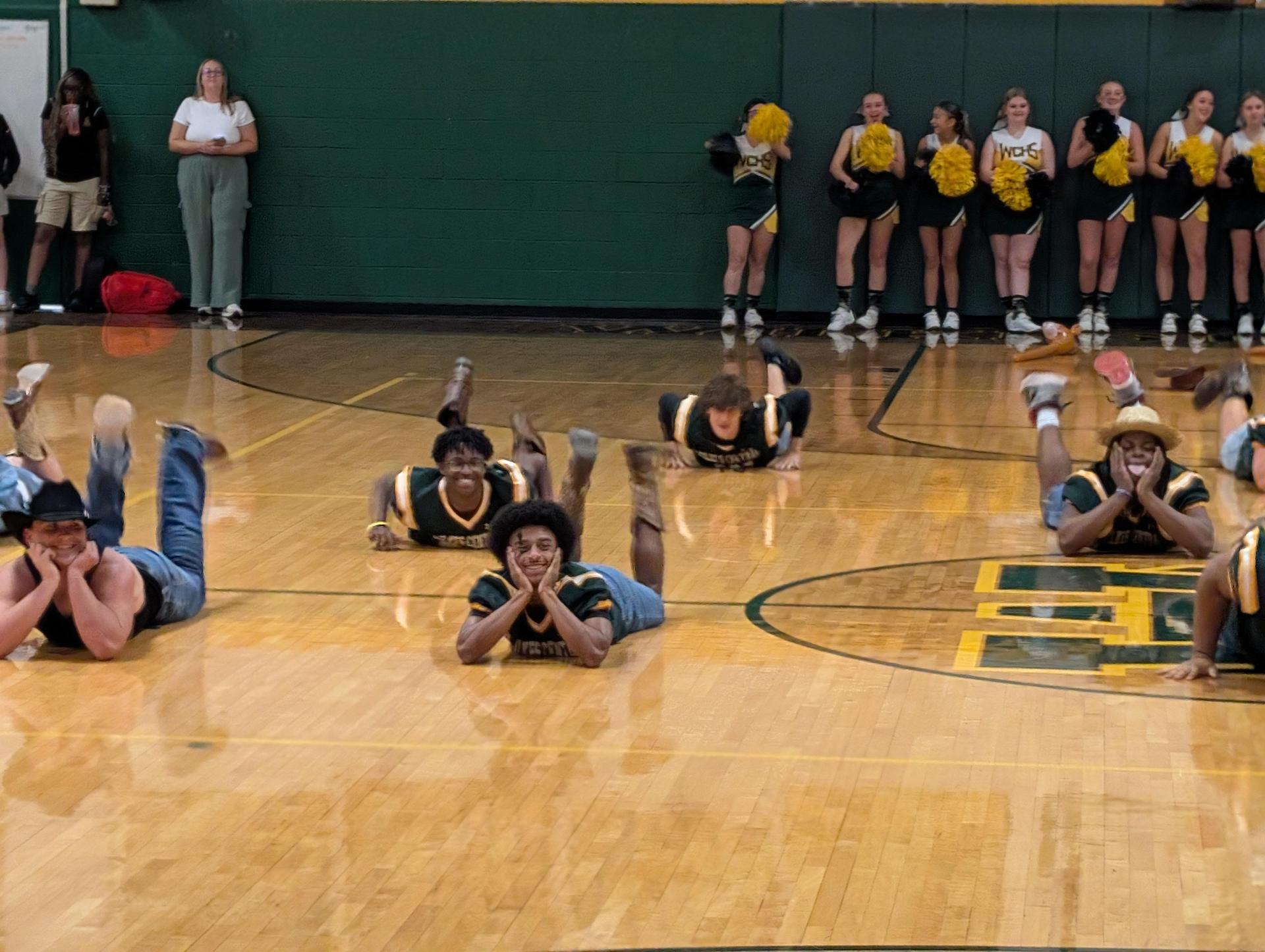Students lying on the gym floor, engaging in a fun performance with cheerleaders in the background.