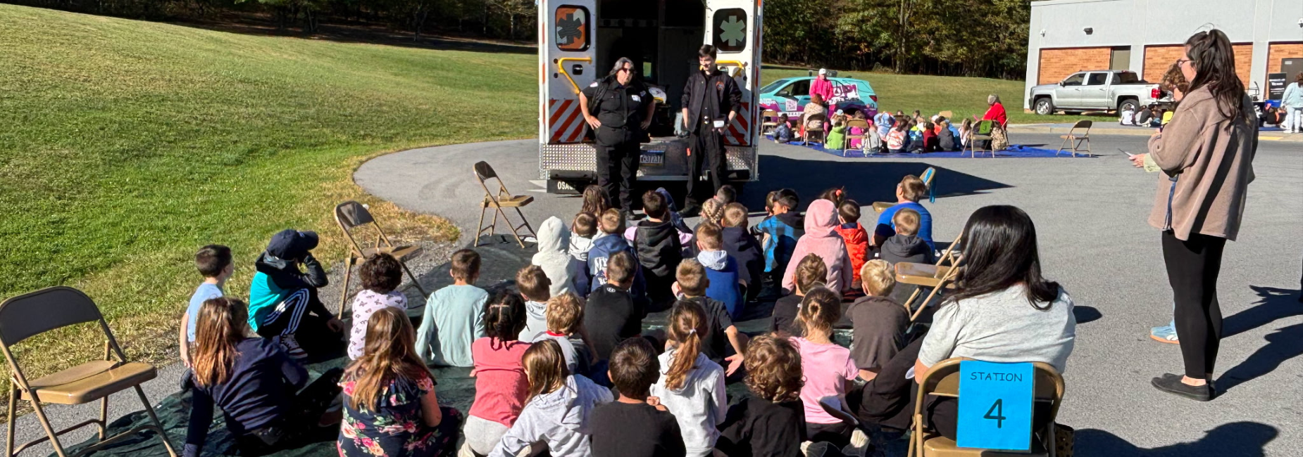 Children seated on a parking lot, listening to first responders by an ambulance.