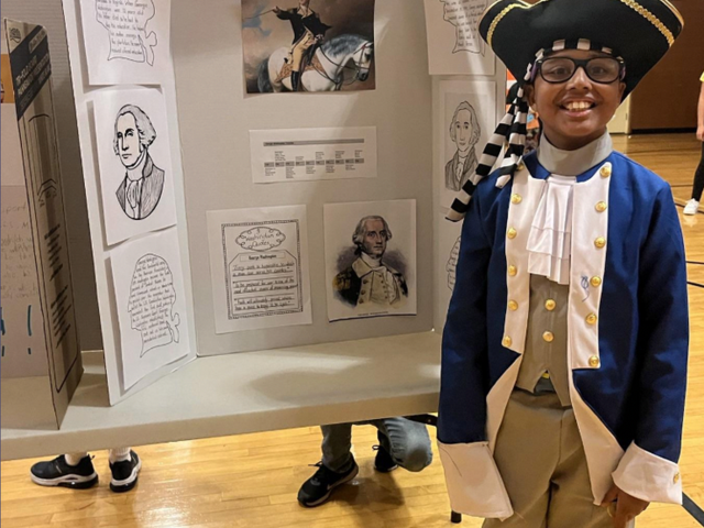 A child dressed as George Washington stands beside a display board with historical illustrations.