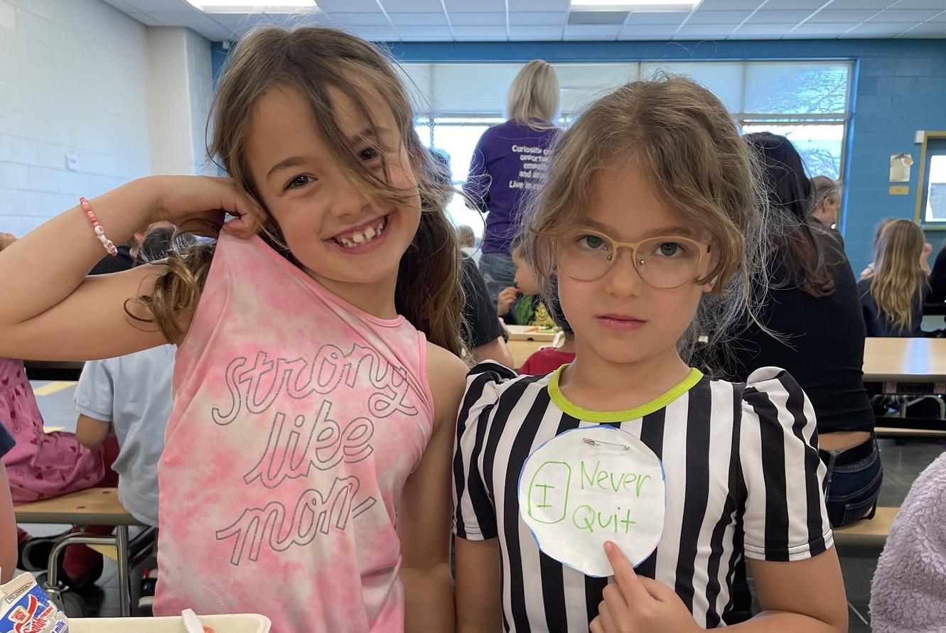 Two girls in a school cafeteria, one wearing a 'Never Quit' sign.