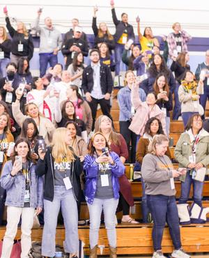 Large crowd of people in a gym, some cheering and holding up phones.