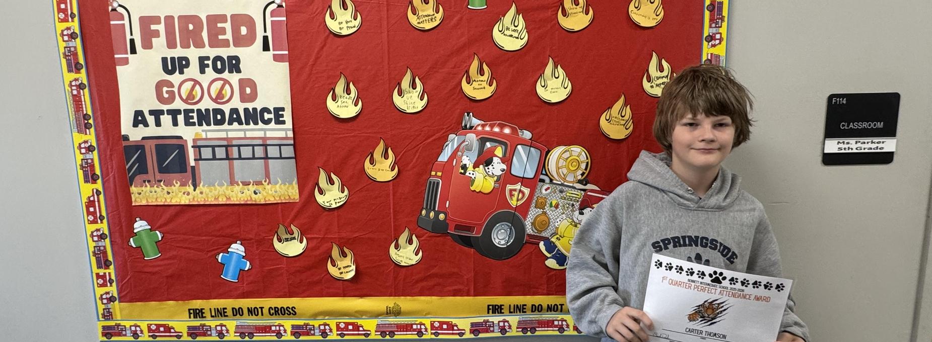 Boy posing with a certificate in front of a fire-themed attendance banner.