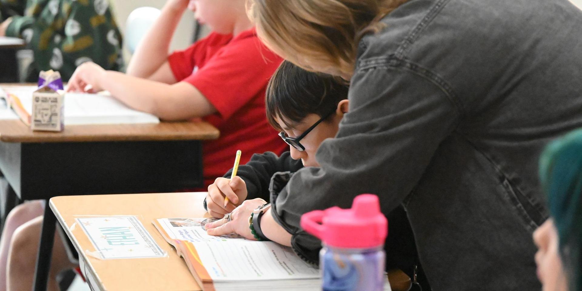 A teacher assists a child with a worksheet at a desk in a classroom.
