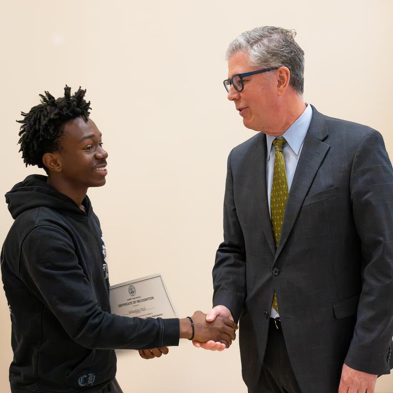 Two men shake hands, one holding a certificate, smiling at each other.