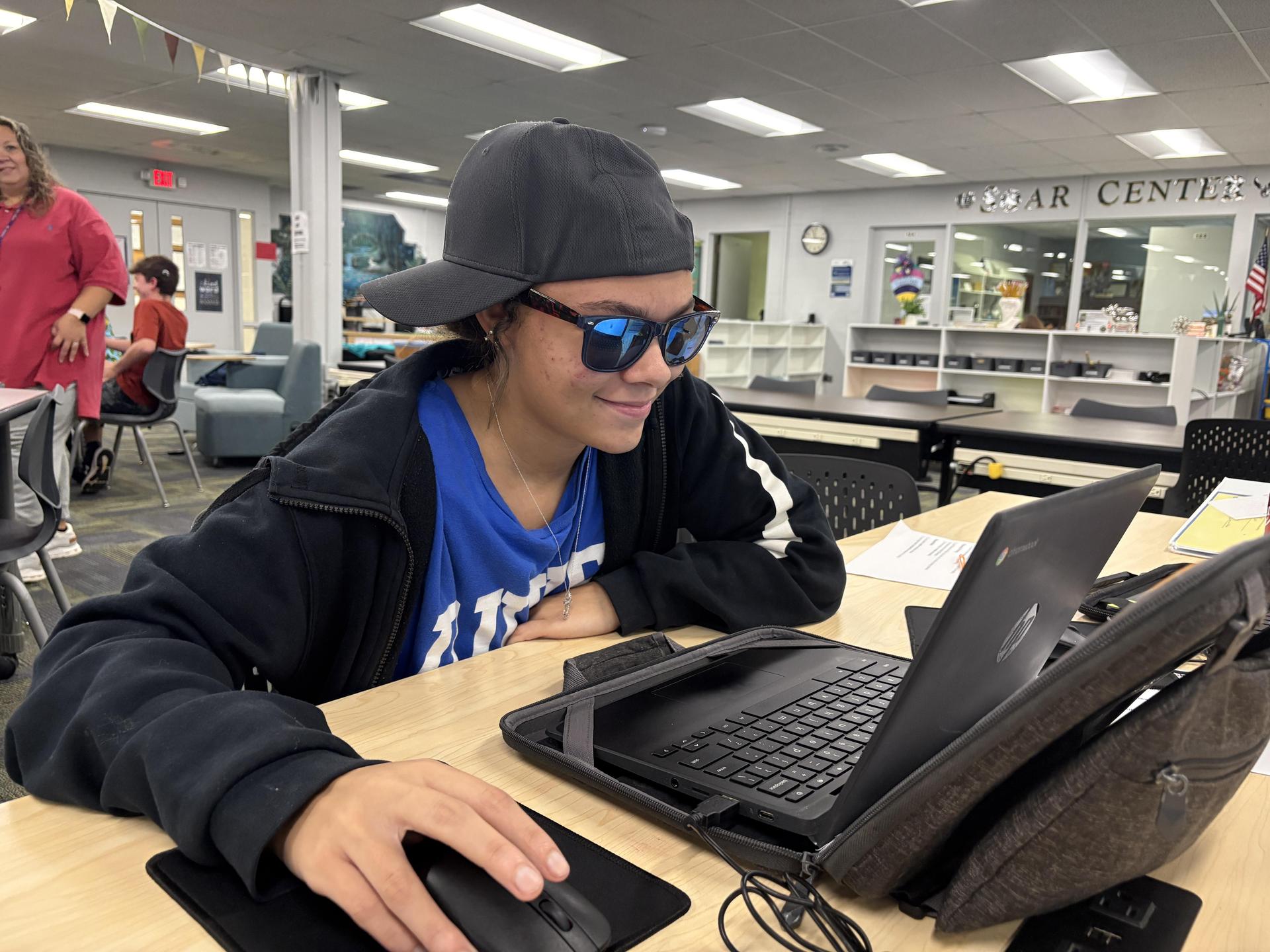Teen wearing sunglasses and a cap smiling at a laptop in a library setting.