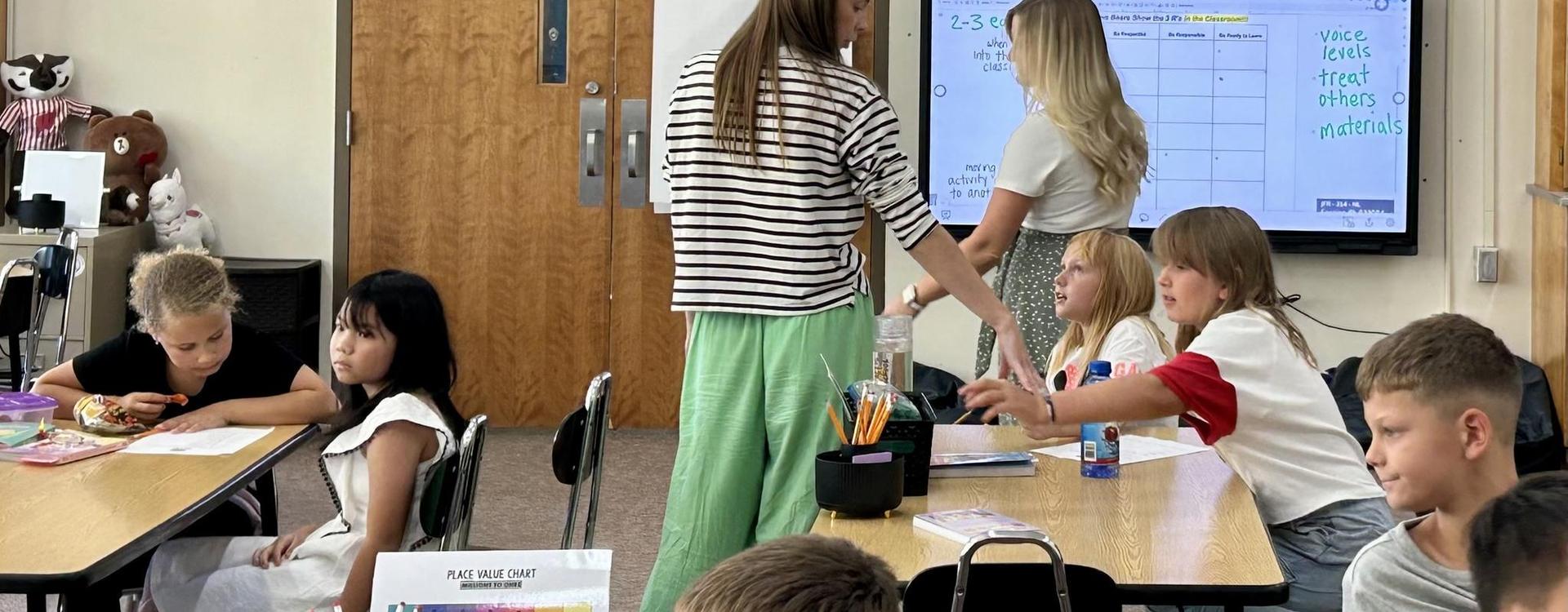 Teacher assisting students at desks while others work on assignments nearby.