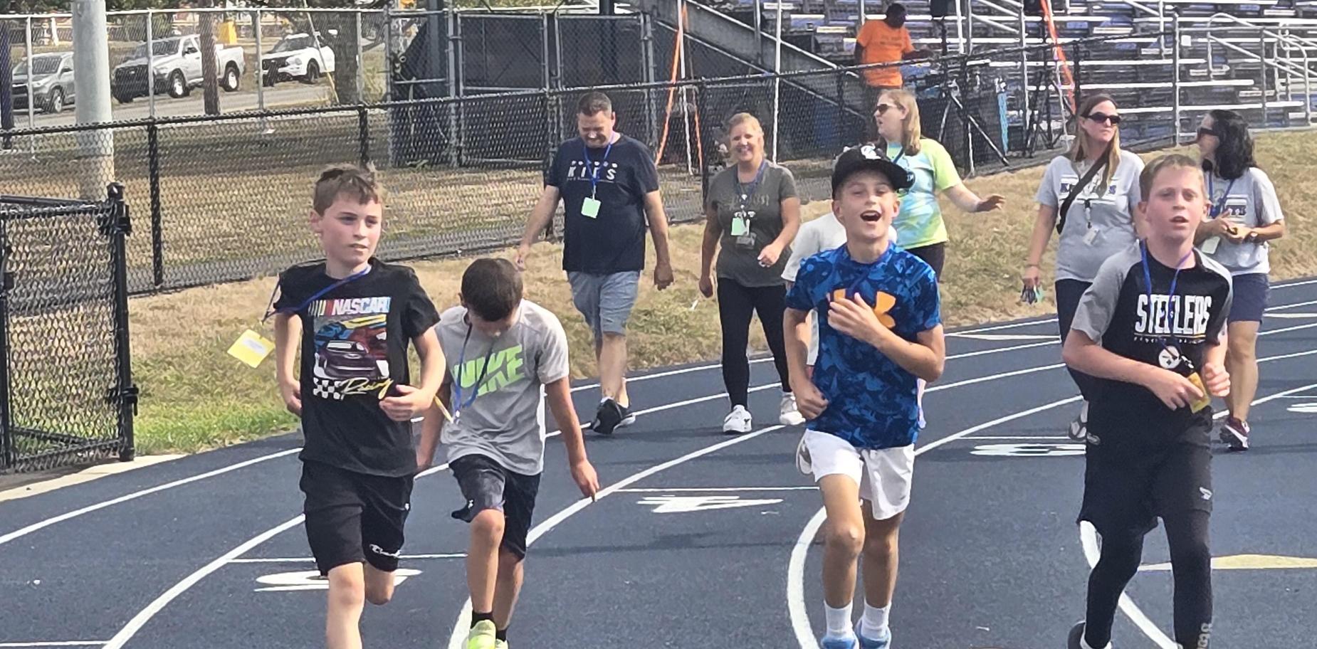 Several boys running together on a track with adults supervising in the background.