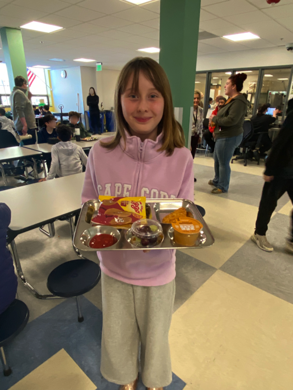 Girl student with short dark blonde hair smiling and holding reusable tray full of food.