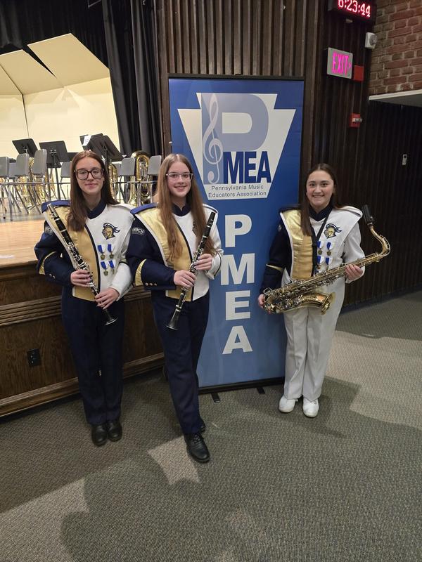 three girl band members in uniform holding their instruments in front of PMEA sign