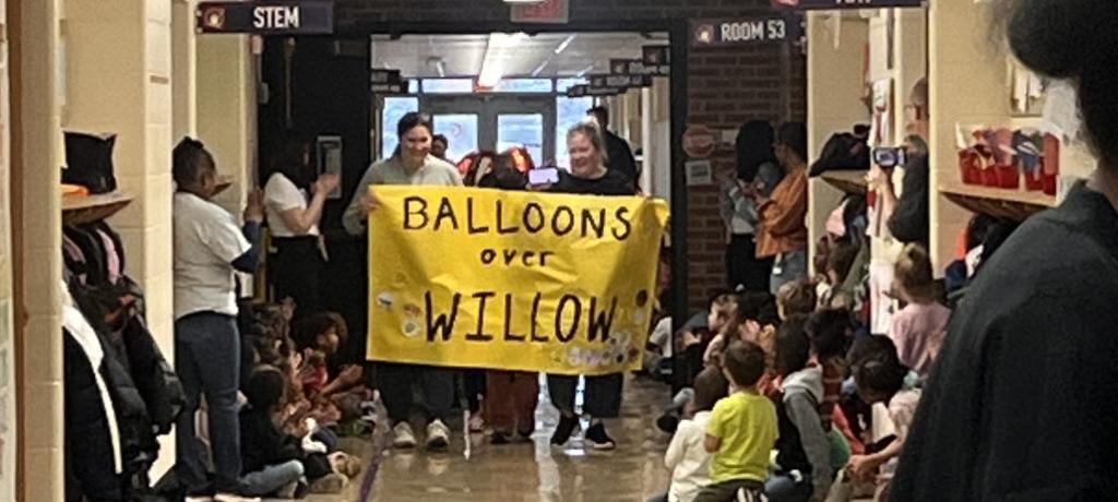 two teachers hold sign that says Balloons Over Willow while students watch along a hallway