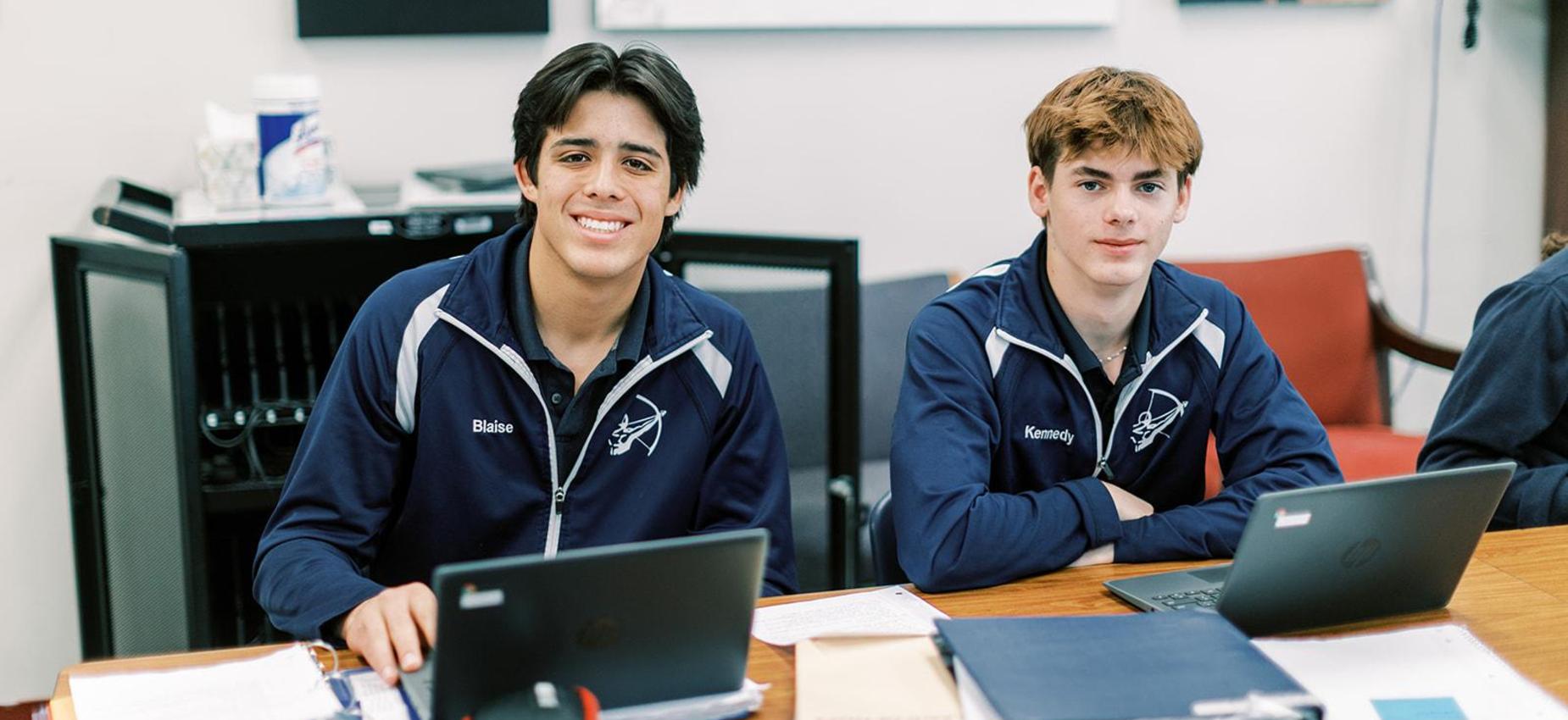 Two smiling high school students in Archers jackets work at their laptops