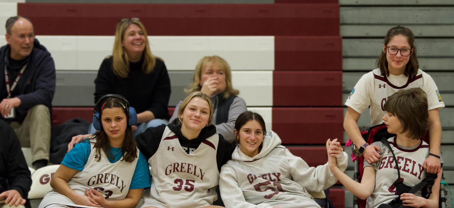 Group of basketball players in team uniforms seated on a bench, smiling and interacting.