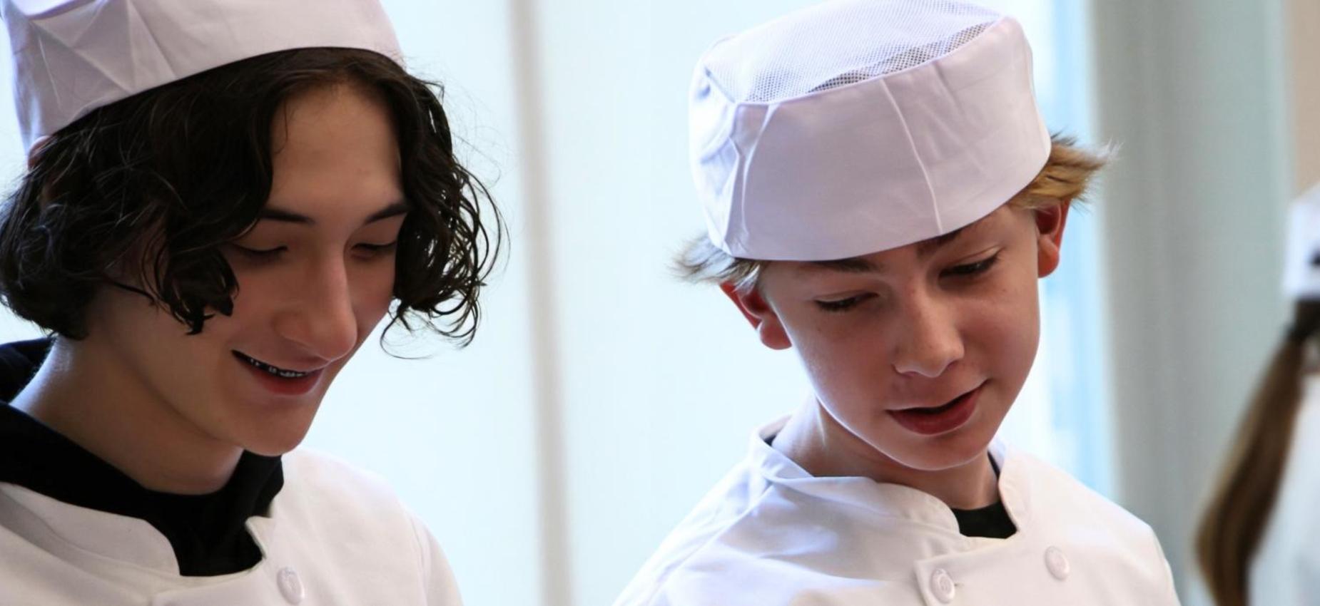 Two boys in chef hats looking at something while cooking together.